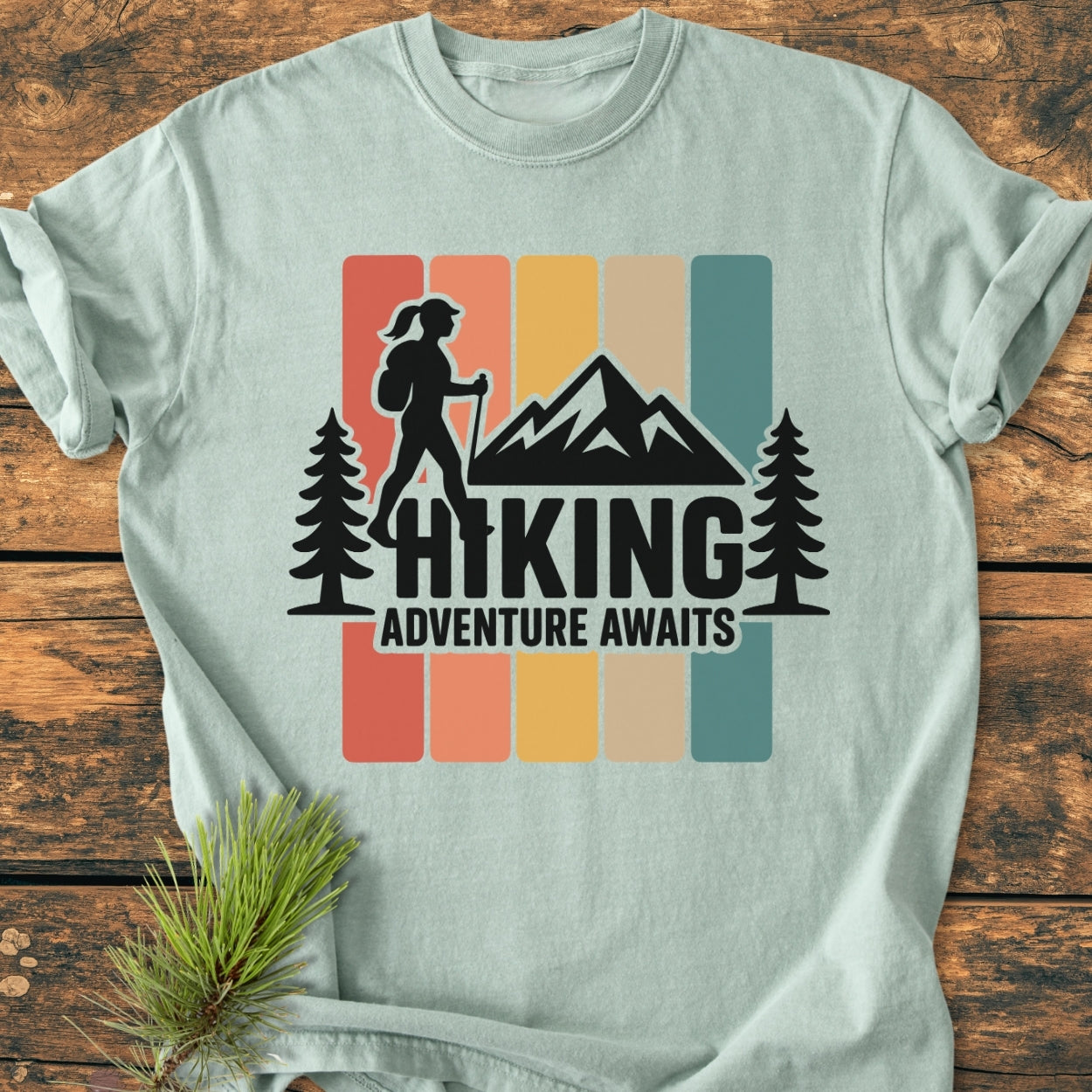 Hiking Girl - Vintage Wash Tee featuring a stylized hiker with trekking pole, flanked by pine trees and mountains, printed on a soft, relaxed-fit, garment-dyed T-shirt laid on a wooden surface.
