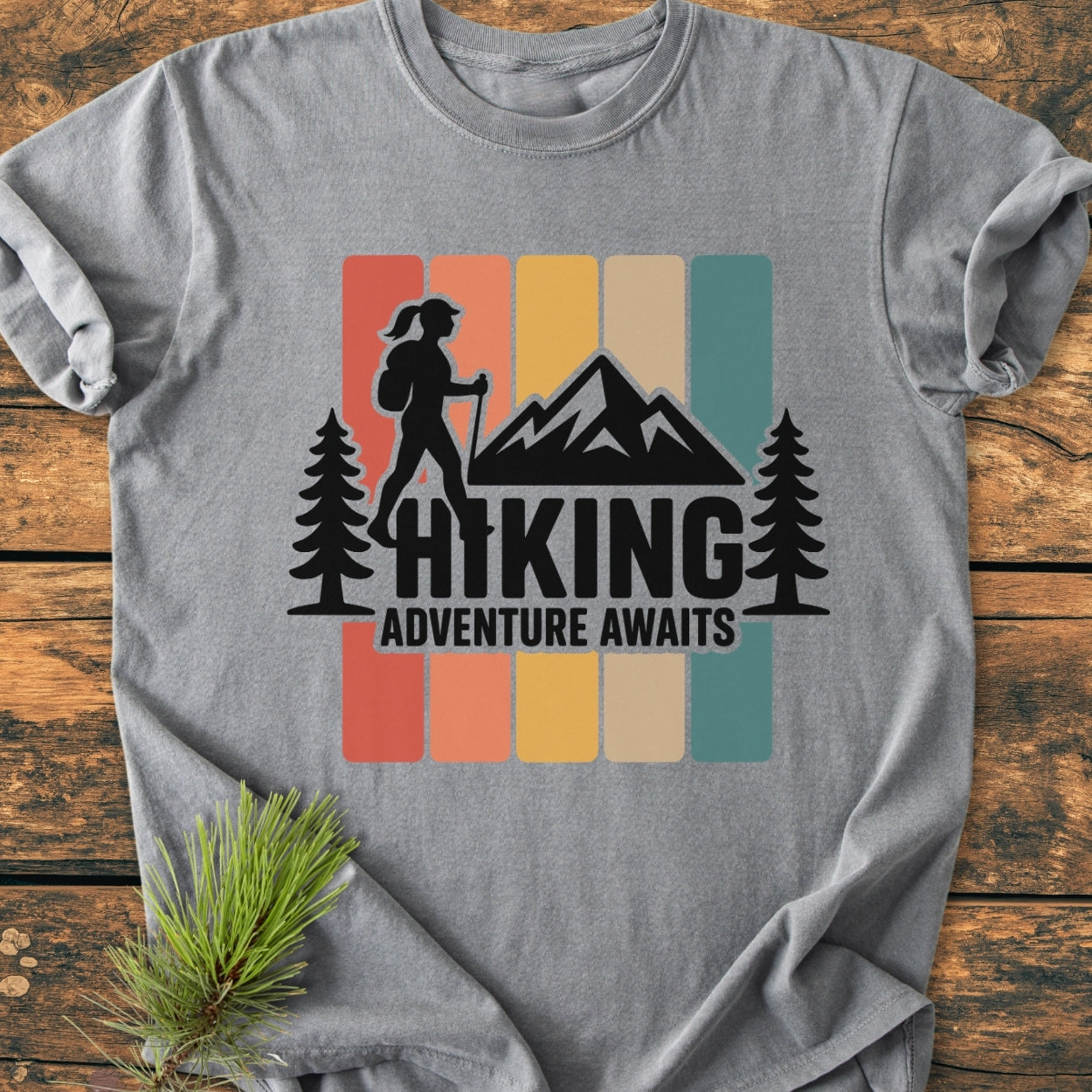 Hiking Girl - Vintage Wash Tee features a retro graphic of a backpacking hiker with pine trees and mountain peak, on a heather-grey T-shirt, laid on a wooden surface, showcasing adventure-inspired design.