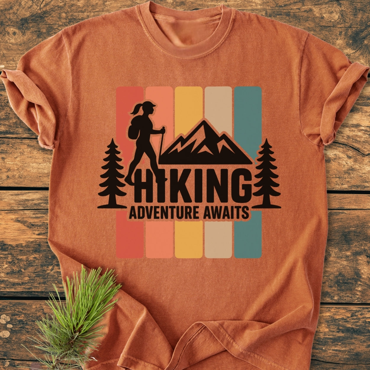 Hiking Girl - Vintage Wash Tee features a hiking scene with a hiker silhouette, trees, and mountain, on a rust-orange backdrop. Displayed on wood, pine needles accent the soft 100% cotton fabric.