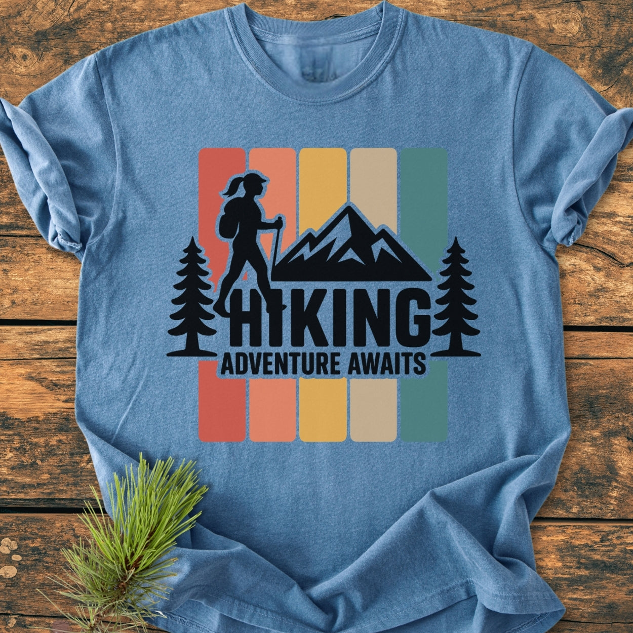 Hiking Girl - Vintage Wash Tee in heather-blue, featuring retro color blocks and a backpacker silhouette with mountain peak. Displayed on wood, includes “HIKING” and “ADVENTURE AWAITS” text, alongside a pine sprig.
