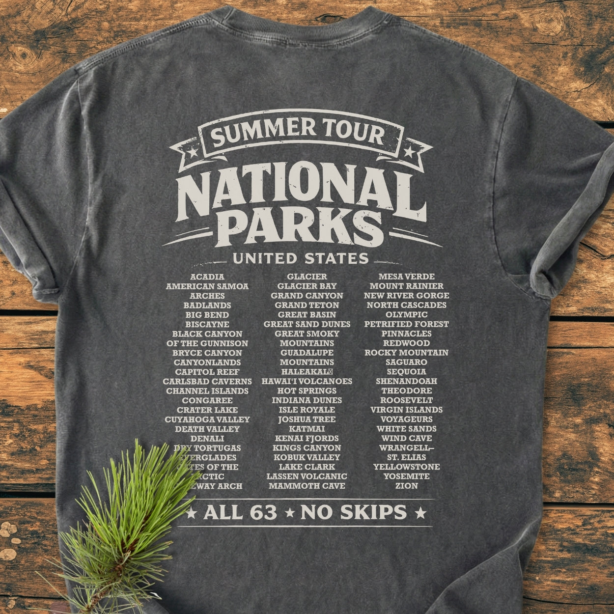 Summer Tour National Parks BACK PRINT - Vintage Wash Tee features a back graphic listing 63 U.S. National Parks, styled on a wooden surface with a pine branch, showcasing its relaxed, durable design.