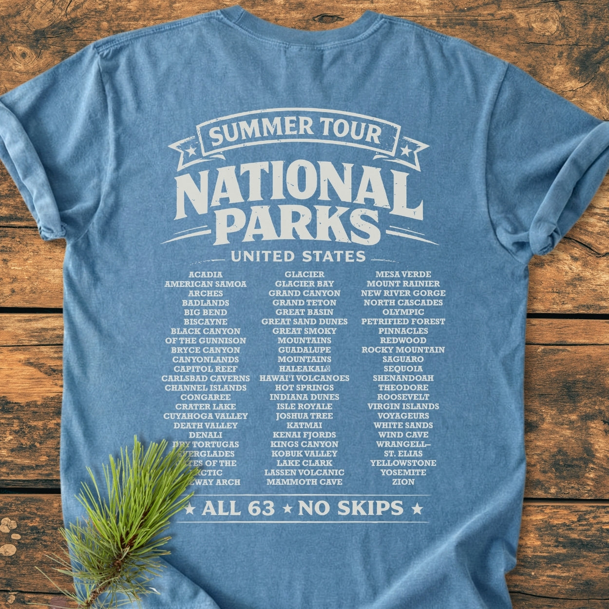 Summer Tour National Parks BACK PRINT - Vintage Wash Tee: Blue T-shirt on wood surface, featuring a 'Summer Tour' design listing all 63 U.S. National Parks on the back, emphasizing 'All 63 · No Skips.'