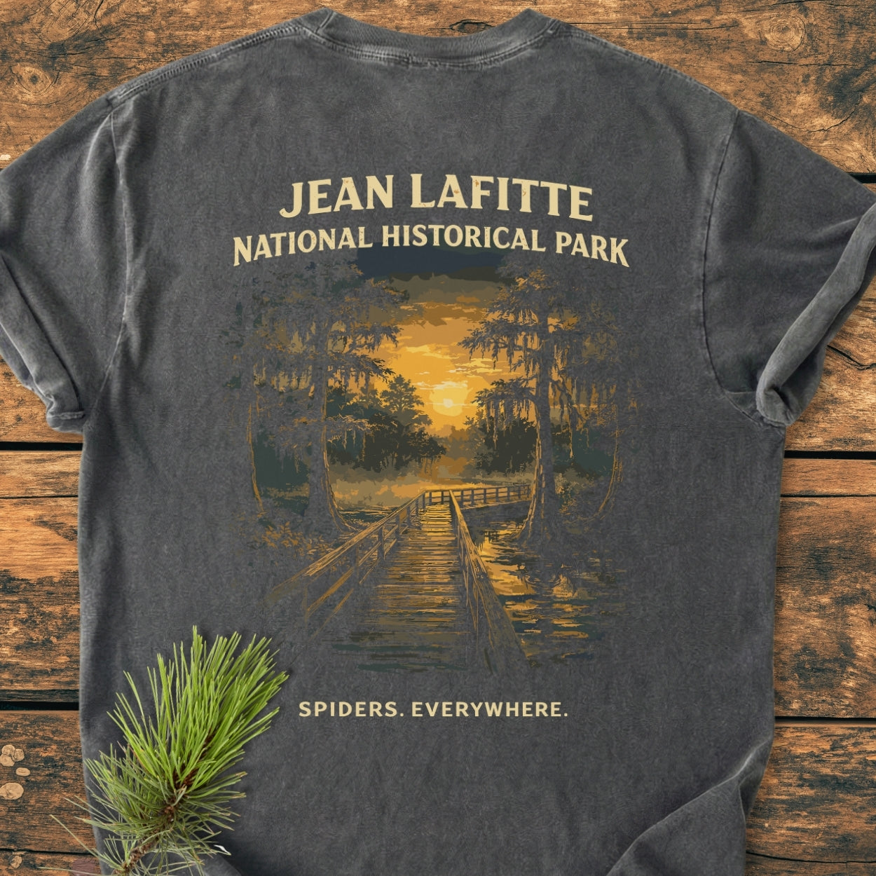 Charcoal-gray Spiders. Everywhere. BACK PRINT - Vintage Wash Tee featuring a sunset boardwalk scene in a cypress swamp, styled with a pine sprig on a wooden surface.