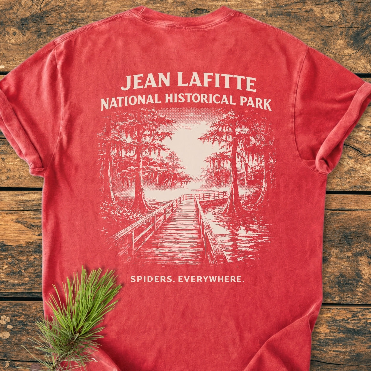 Red T-shirt with JEAN LAFITTE NATIONAL HISTORICAL PARK and SPIDERS. EVERYWHERE. printed on the back, featuring a swamp boardwalk sketch. Laid on wood with a pine sprig.