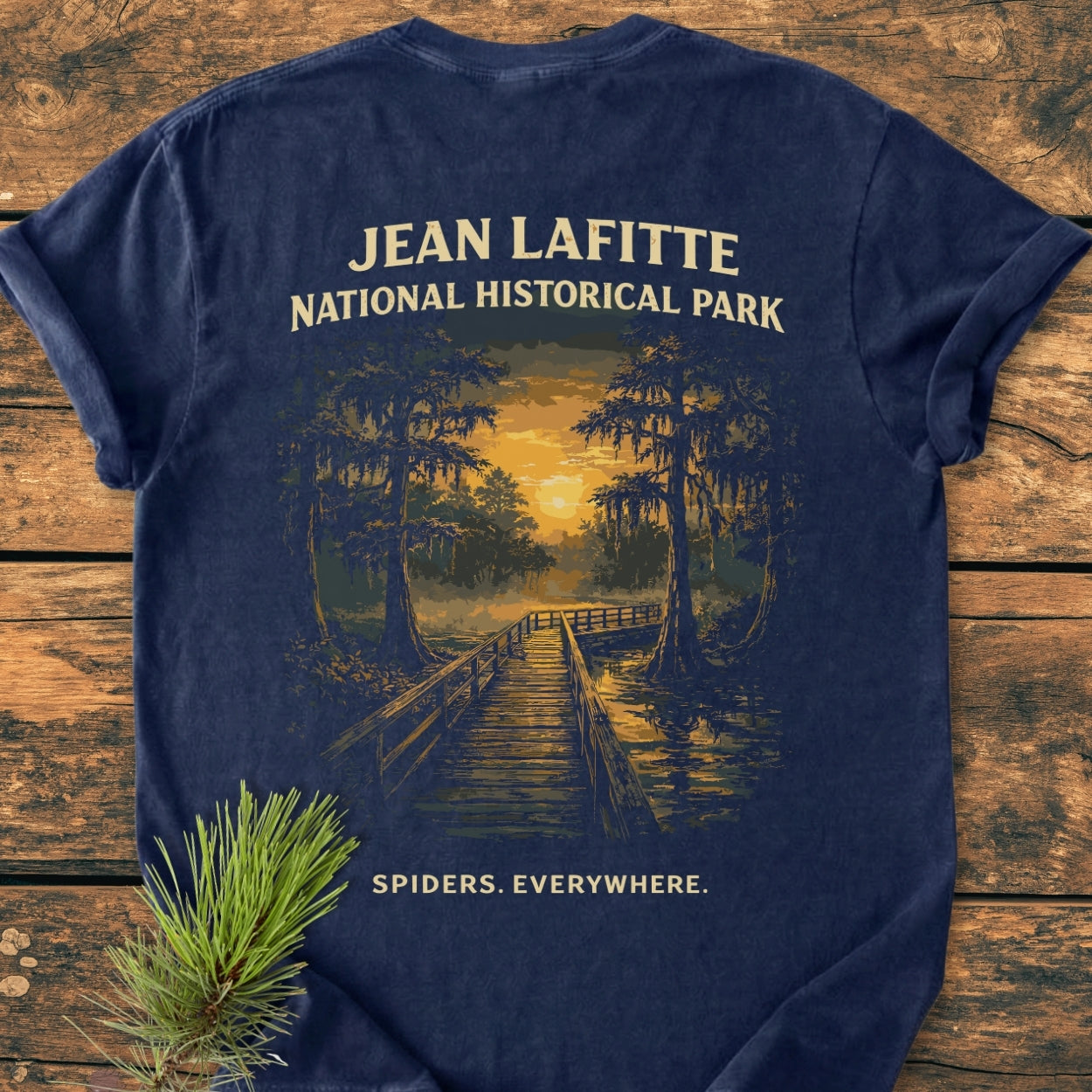 Navy-blue tee showcasing a stylized cypress swamp sunset and boardwalk scene with JEAN LAFITTE NATIONAL HISTORICAL PARK and SPIDERS. EVERYWHERE. text. Laid on wood, pine sprig nearby.