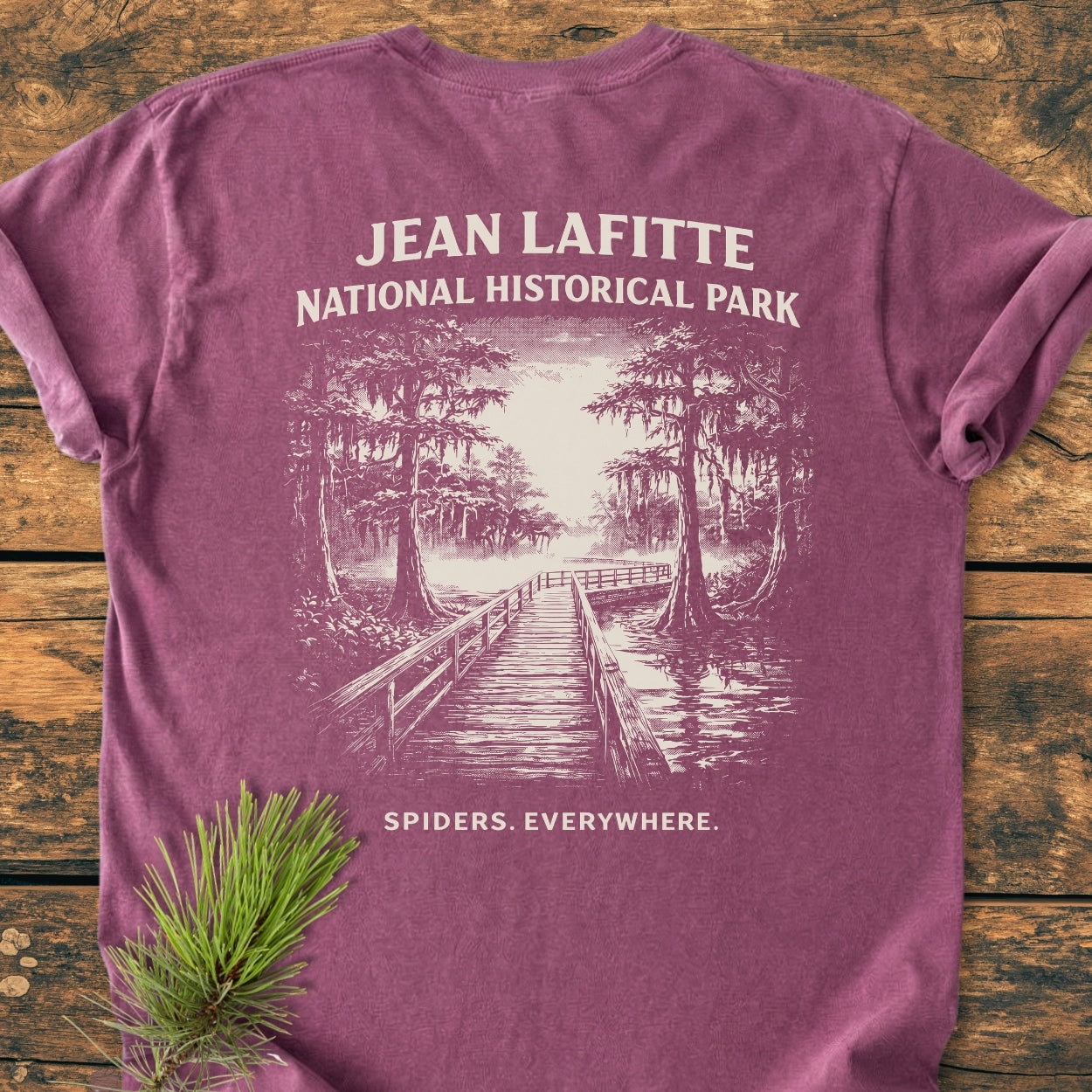 Flat-lay of the Spiders. Everywhere. BACK PRINT - Vintage Wash Tee featuring a swamp scene with cypress trees and tagline, alongside a pine sprig on a wooden surface.