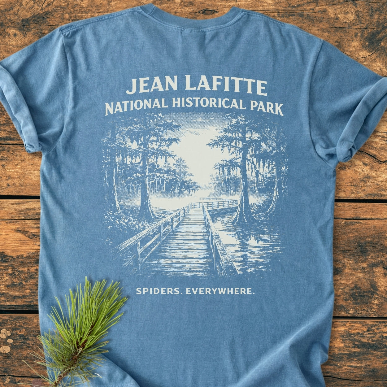 Light-blue tee titled Spiders. Everywhere. BACK PRINT - Vintage Wash Tee featuring a swamp boardwalk illustration with cypress trees and quirky text, laid on wood with a pine branch beside it.