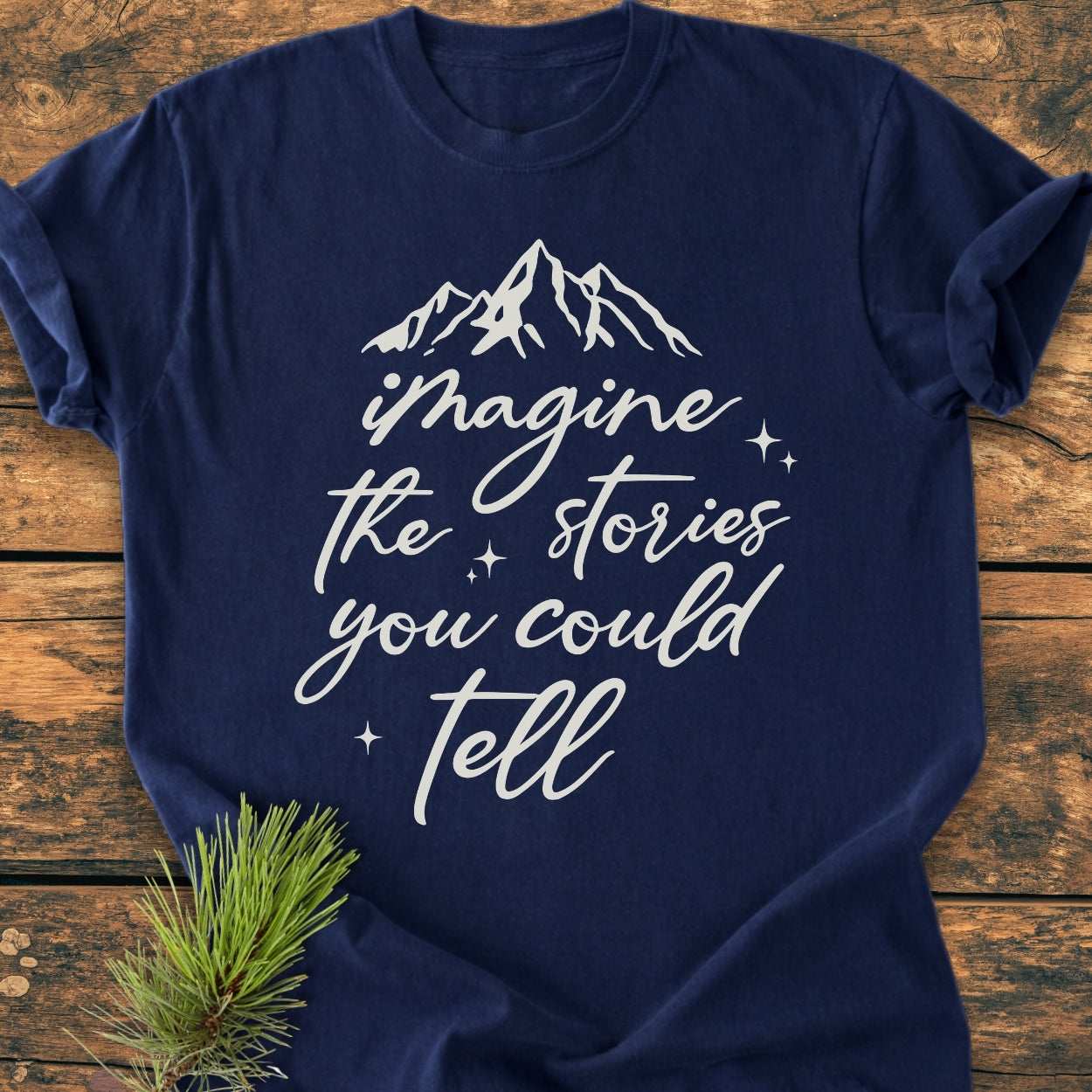 Imagine The Stories - Vintage Wash Tee, a navy-blue shirt with a mountain silhouette and slogan in white, surrounded by star accents, laid flat on a wooden surface with a pine twig beside it.
