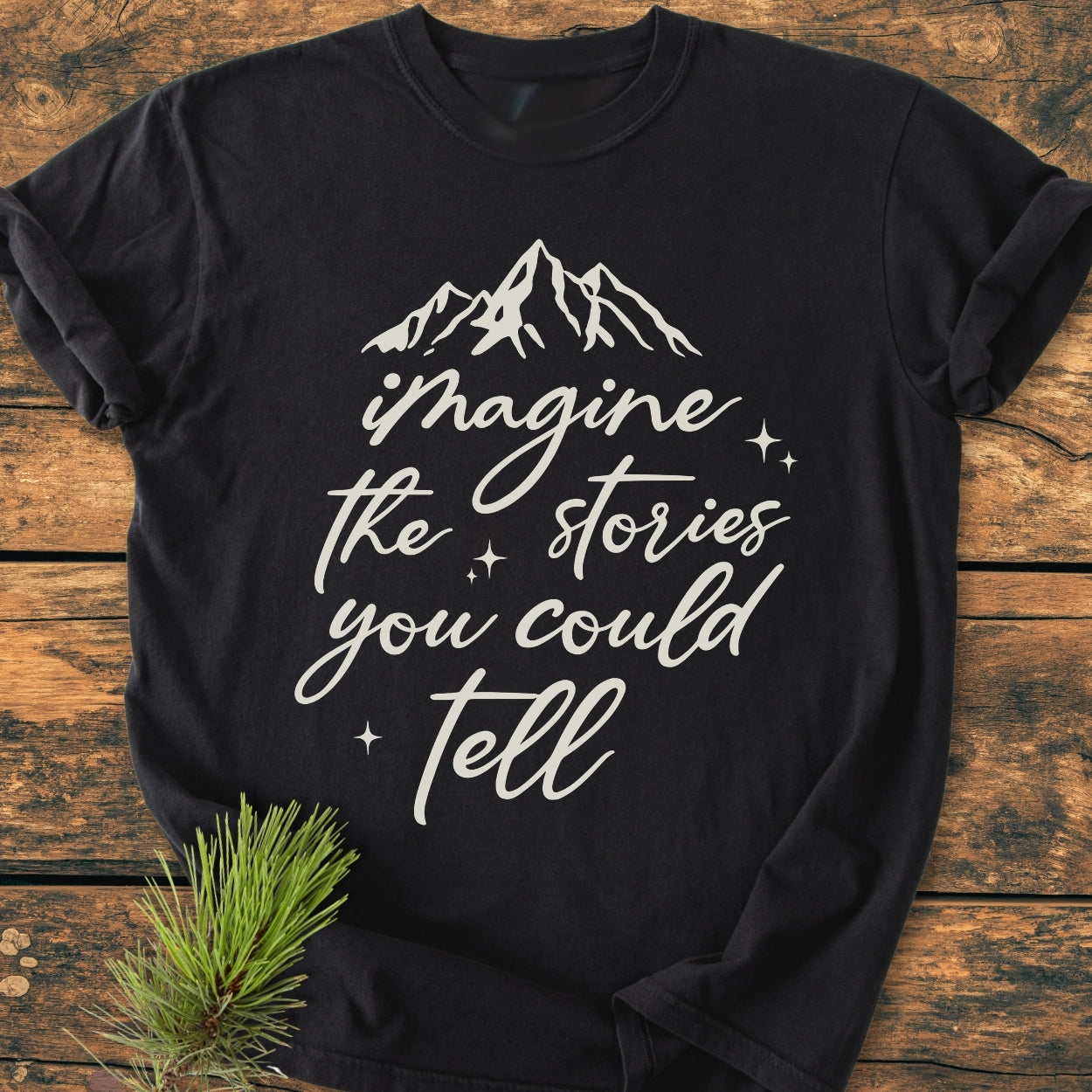 Imagine The Stories - Vintage Wash Tee with mountain graphic and text, laid flat on wood. Features a soft, relaxed fit, pre-shrunk 100% ring-spun cotton. Pine sprig adds natural touch.