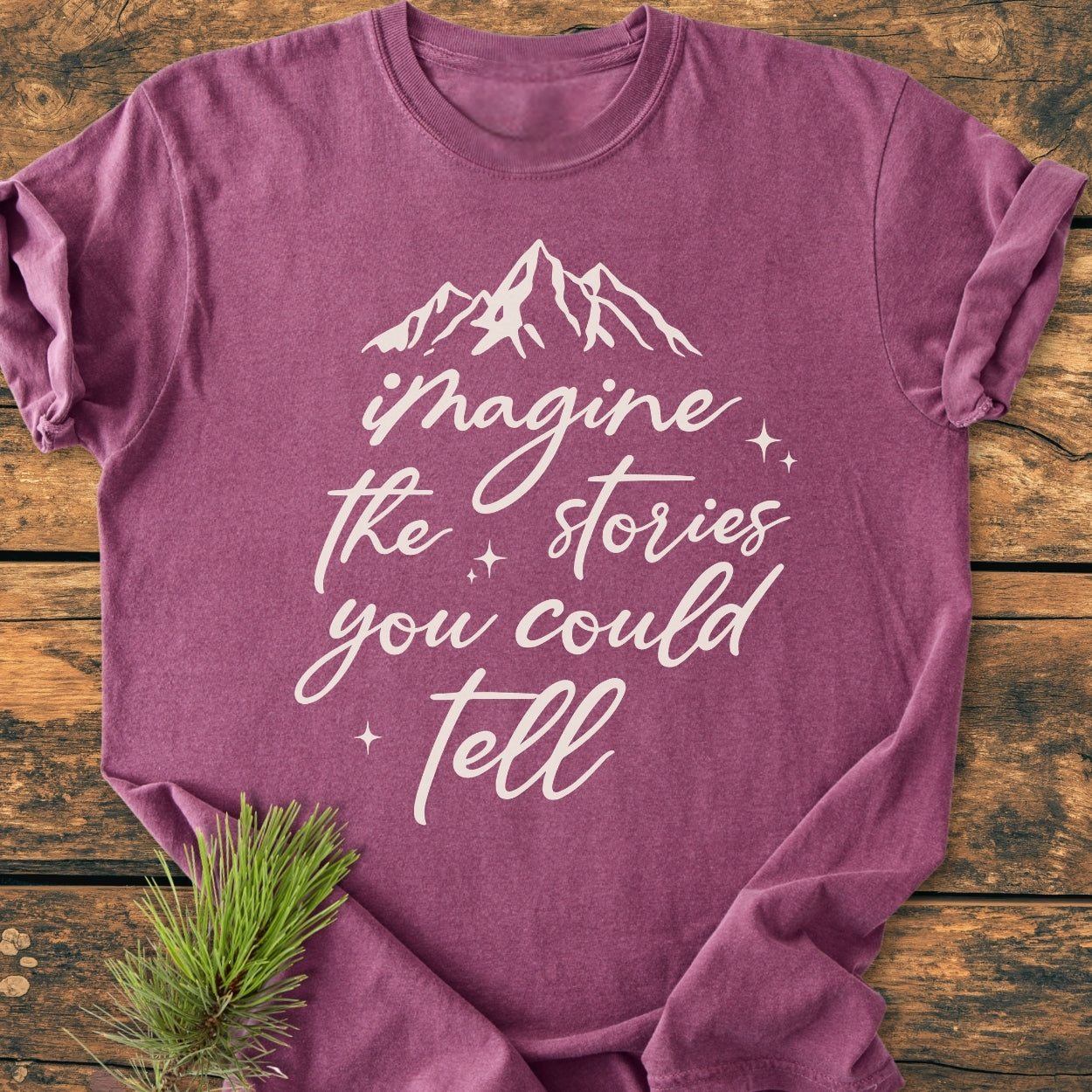 Imagine The Stories - Vintage Wash Tee with white script and mountain silhouette, laid on wood. Features a cozy, relaxed fit, pre-shrunk ring-spun cotton, showcasing durability with double-needle stitching.