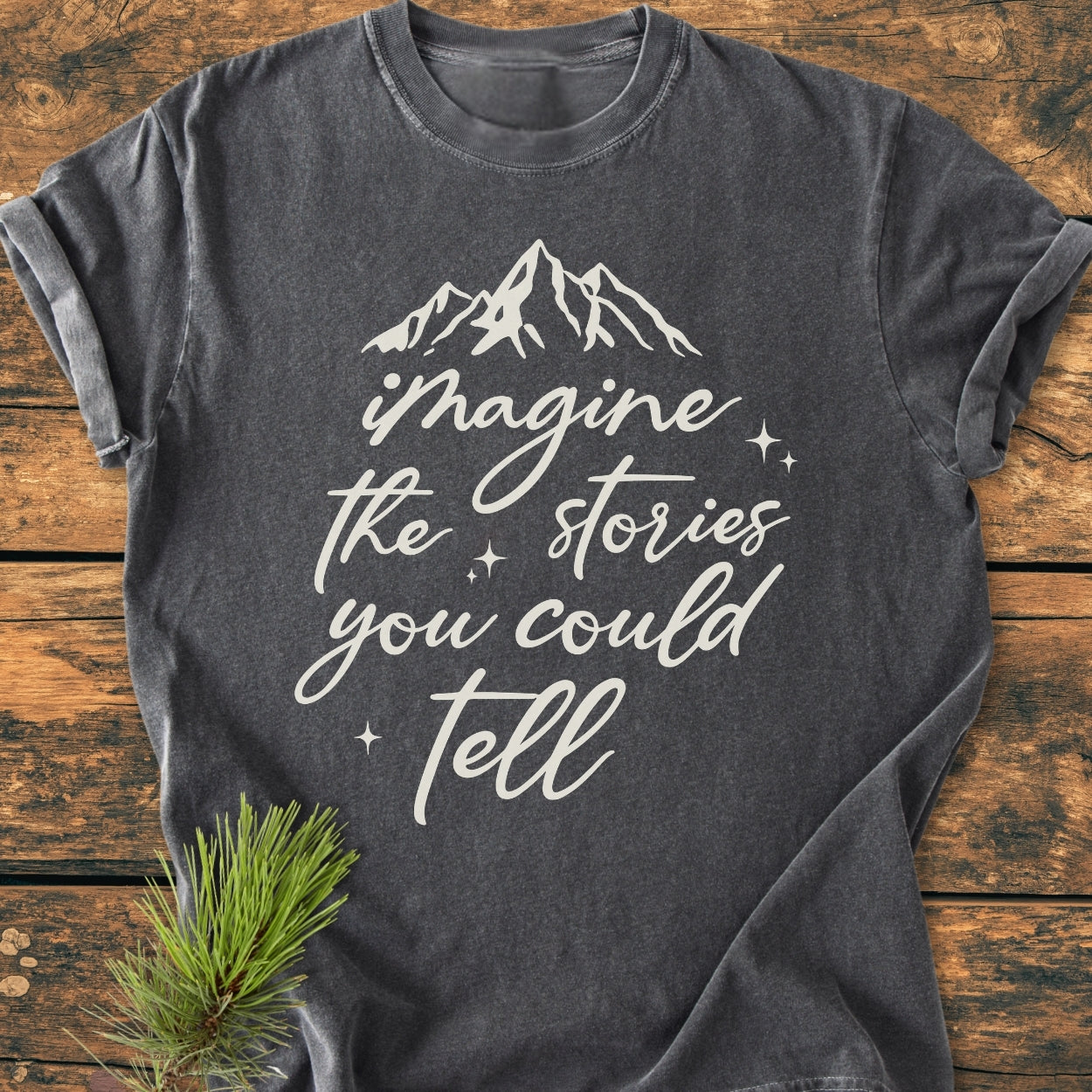 Imagine The Stories - Vintage Wash Tee in dark charcoal-gray with white script and mountain motif, displayed on a wooden surface with a pine sprig accent, showcasing its relaxed, cozy fit.
