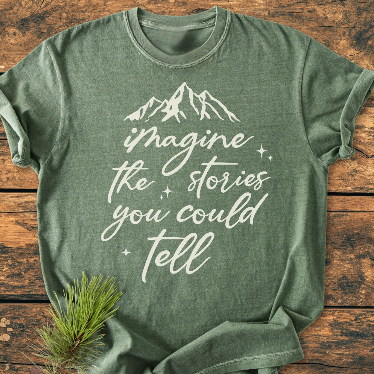 Imagine The Stories - Vintage Wash Tee: Heather-green T-shirt with a mountain range and phrase graphic, laid flat on wood. Crafted from 100% ring-spun cotton for comfort and durability, featuring a relaxed fit.