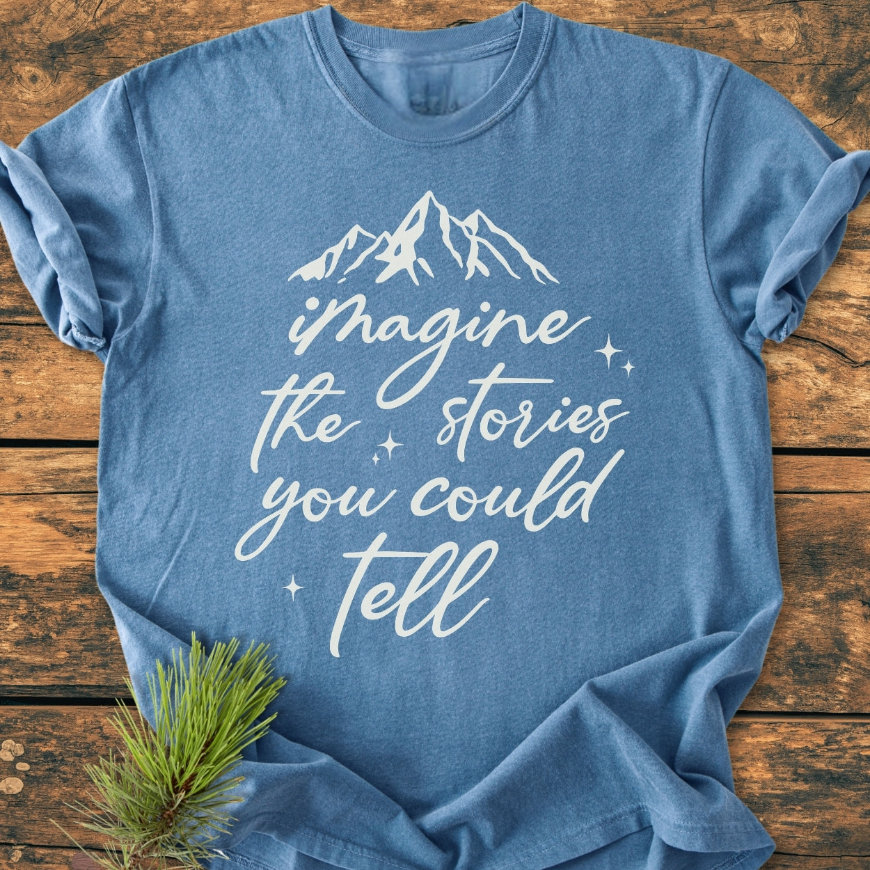 Imagine The Stories - Vintage Wash Tee features a heather-blue color with a white mountain graphic and slogan. Laid flat on wood, it includes a sprig of pine, highlighting its rustic charm.