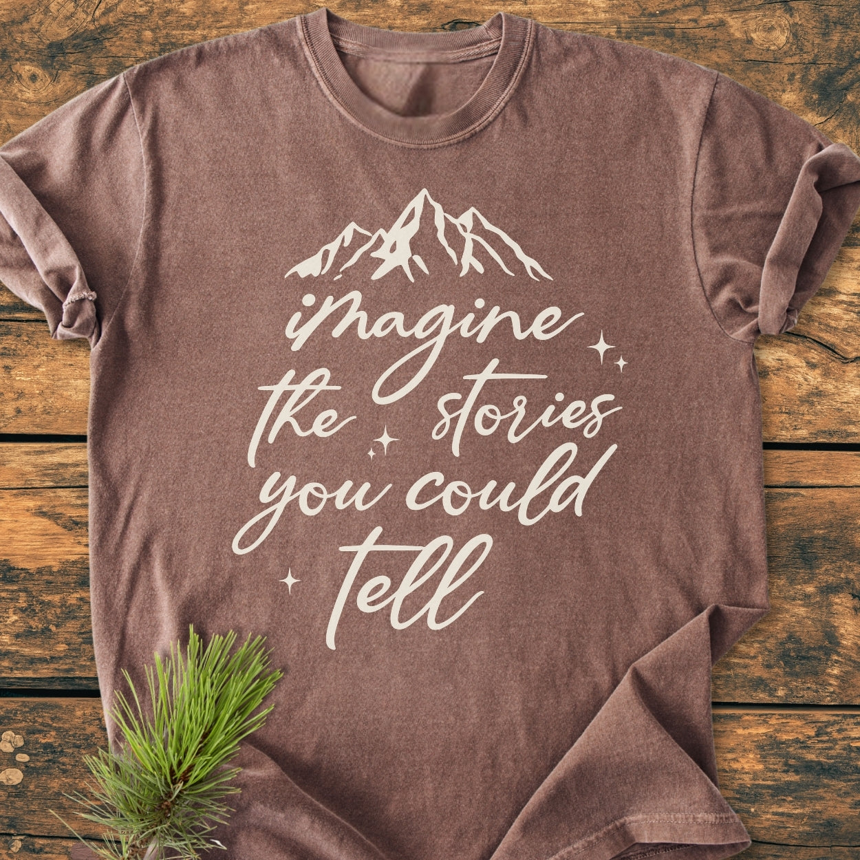 Imagine The Stories - Vintage Wash Tee features a brown cotton T-shirt with a mountain silhouette and phrase, laid on wood with a pine sprig. Crafted from 100% ring-spun cotton for comfort and durability.