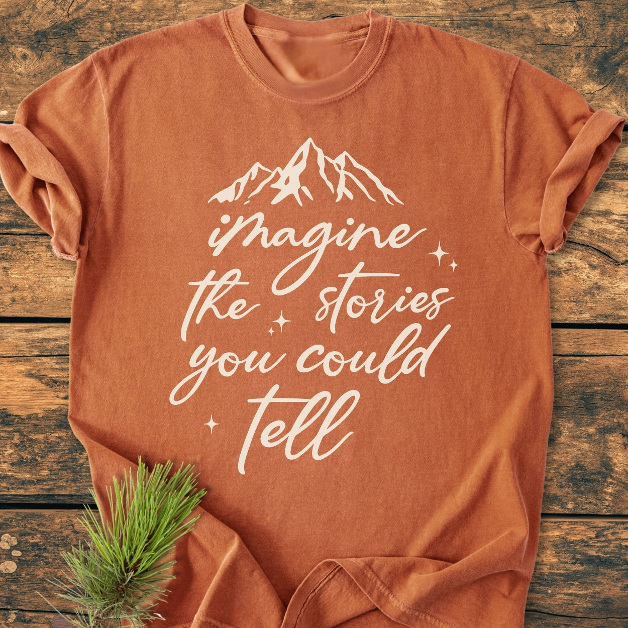 Imagine The Stories - Vintage Wash Tee folded on rustic wood, featuring a white mountain graphic and script slogan. A pine sprig enhances its outdoorsy vibe, highlighting the tee's cozy, relaxed style.