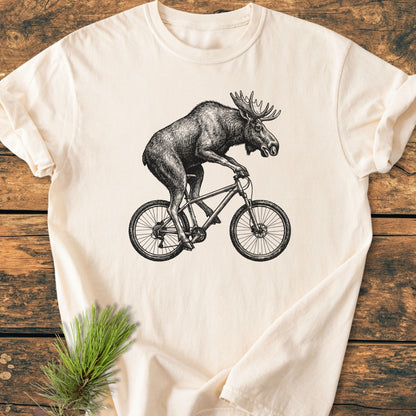 Moose On The Road - Vintage Wash Tee featuring a monochrome moose on a bicycle illustration, laid on a wooden surface with a pine sprig beside it, highlighting its relaxed fit and durable stitching.