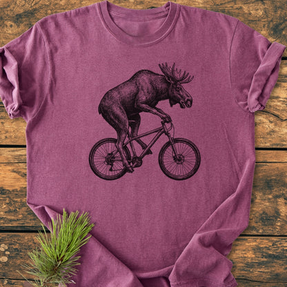 Moose On The Road - Vintage Wash Tee featuring a black moose on a bicycle line-drawing, laid on wood with a pine sprig. Crafted from 100% ring-spun cotton for a cozy, relaxed fit.