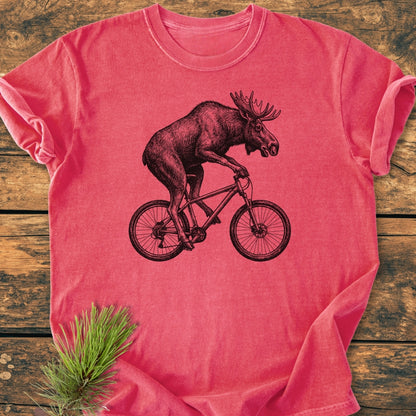 Moose On The Road - Vintage Wash Tee features a sketch of a moose cycling, printed on a soft, heathered-red tee. Laid on wood, it includes a decorative pine branch.