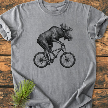 Moose On The Road - Vintage Wash Tee: Heather-gray T-shirt with a black line drawing of a moose on a bicycle, laid on wood. A pine sprig adds a rustic touch.