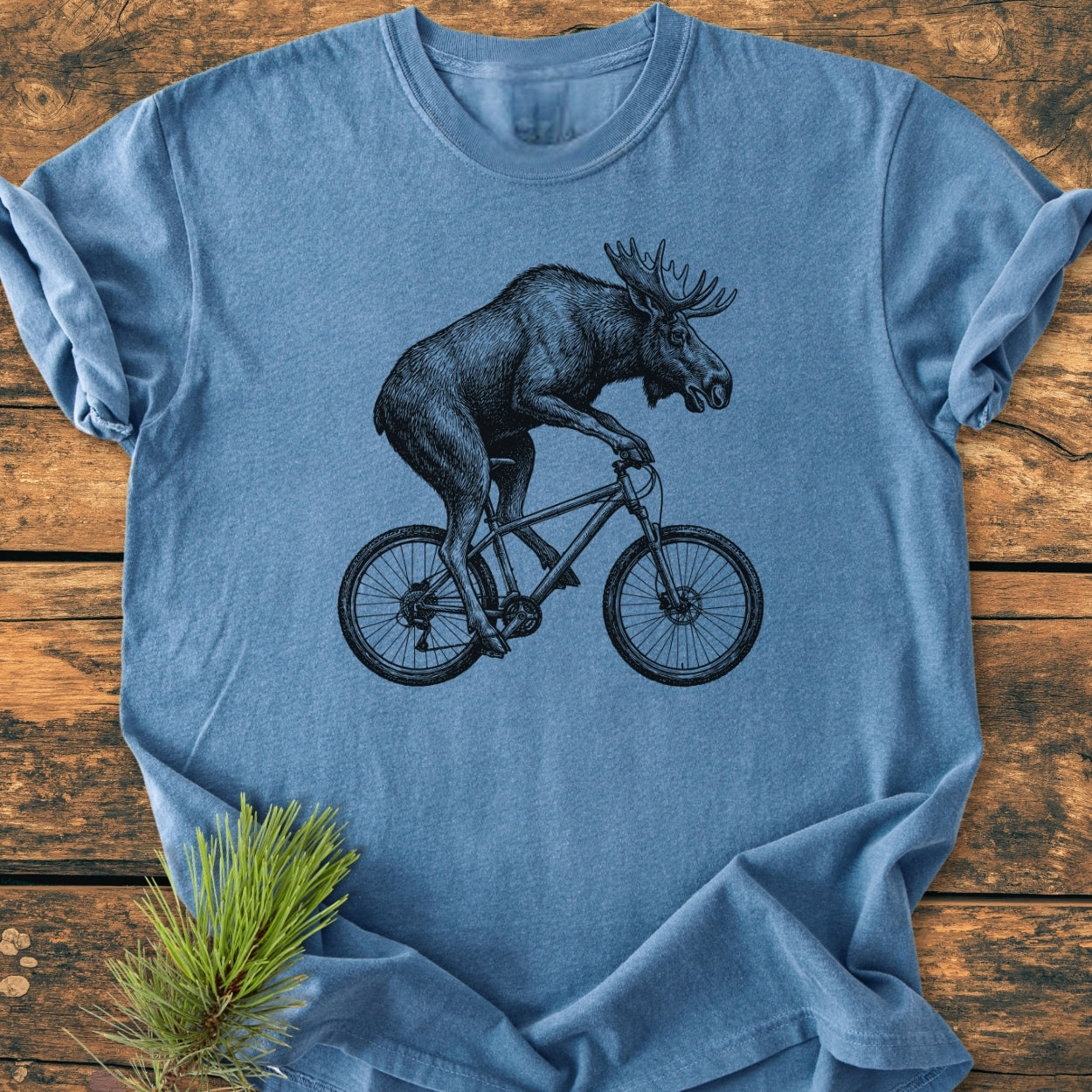 Moose On The Road - Vintage Wash Tee featuring a black-ink illustration of a moose cycling, laid flat on a wooden surface with pine branch. Heather-blue, relaxed fit, 100% ring-spun cotton.