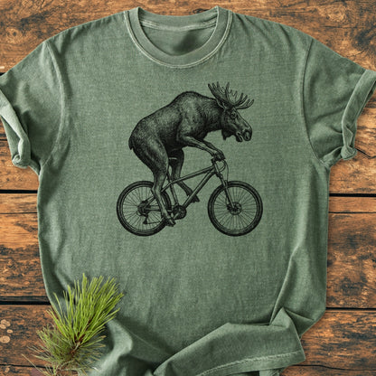 Moose On The Road - Vintage Wash Tee: Heather-green tee with a moose riding a bicycle design, displayed on wood with pine sprig, showcasing relaxed fit and durable stitching for daily wear.