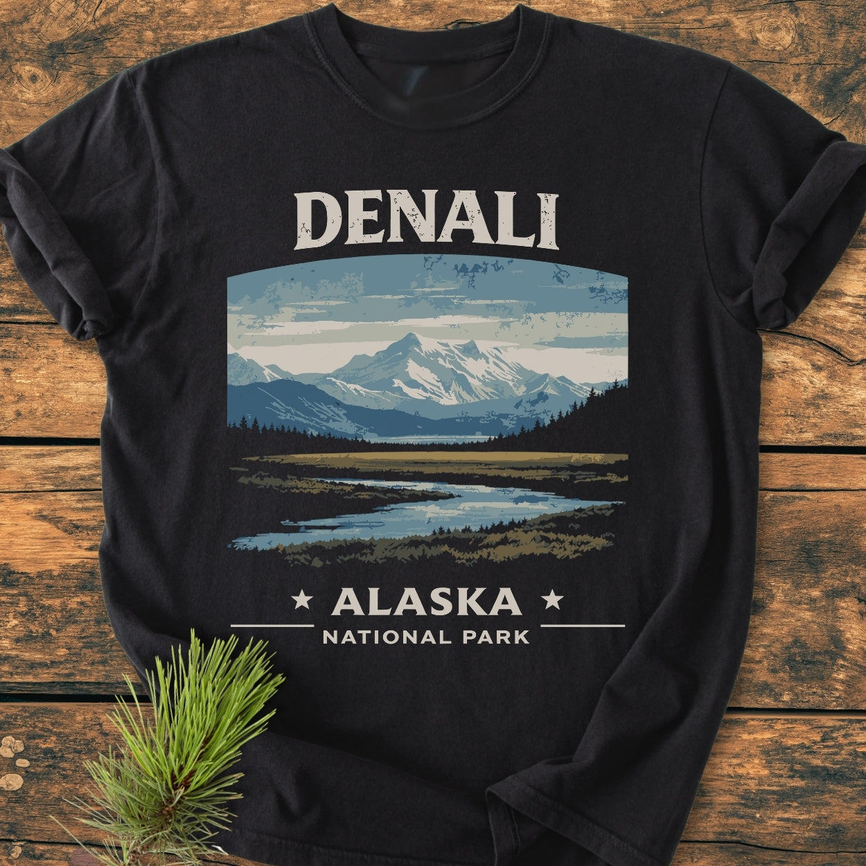Denali National Park - Vintage Wash Tee features a mountain landscape print on a black T-shirt, laid on wood with a pine sprig beside it, highlighting the shirt's durable, relaxed fit design.