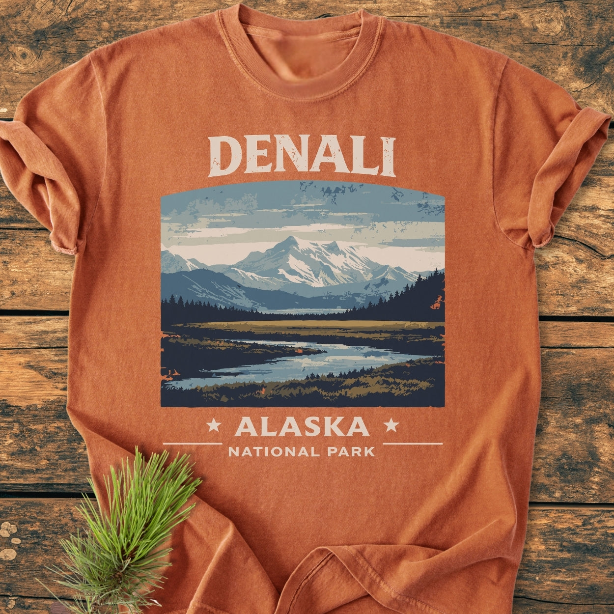 Denali National Park - Vintage Wash Tee featuring a stylized landscape of Denali, with snow-capped mountains, forest, and reflective water, laid flat with a pine sprig accentuating the outdoor theme.