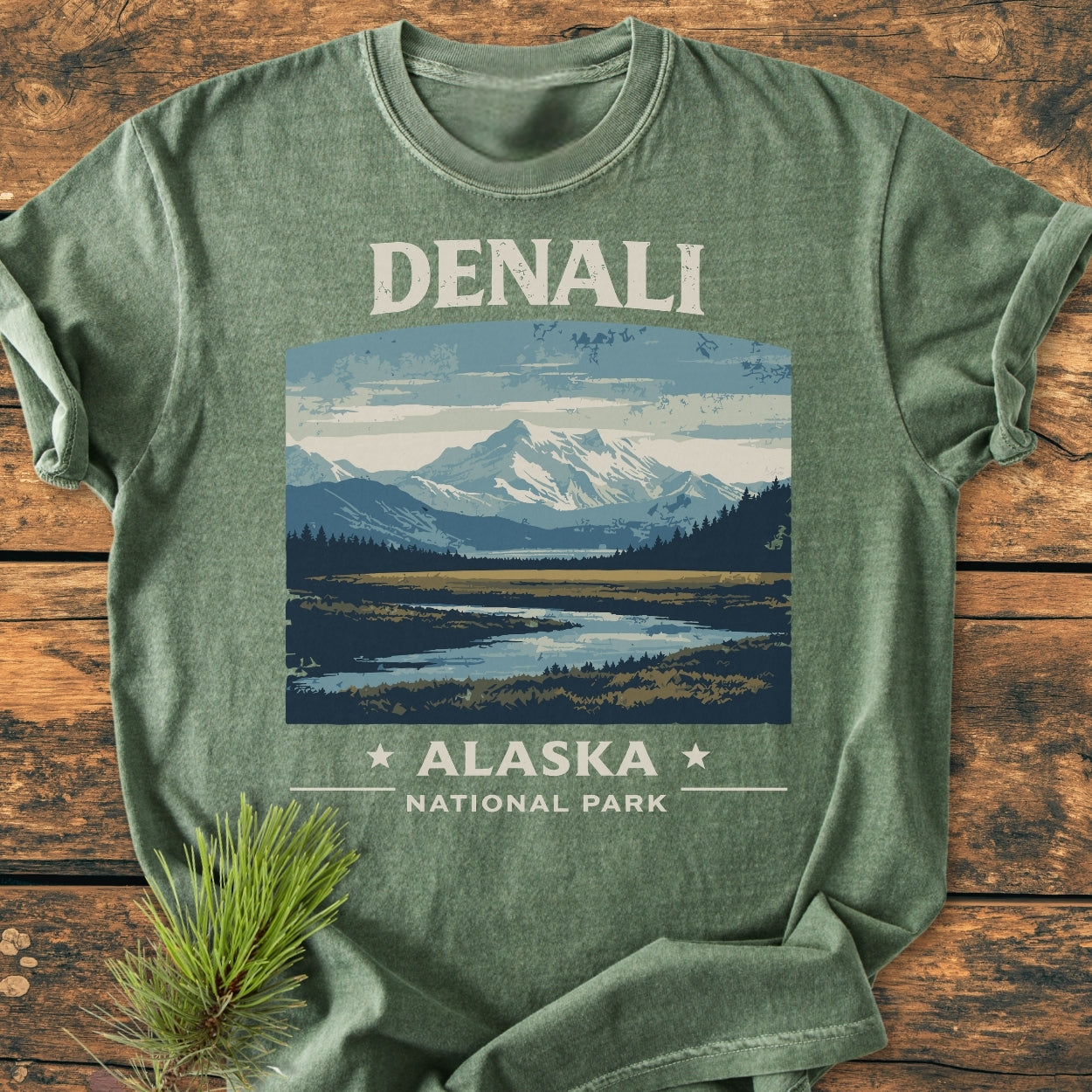 Denali National Park - Vintage Wash Tee featuring snow-capped mountains and treeline on a forest-green background, laid on wood with pine needles. Made from soft-washed, ring-spun cotton for comfort.