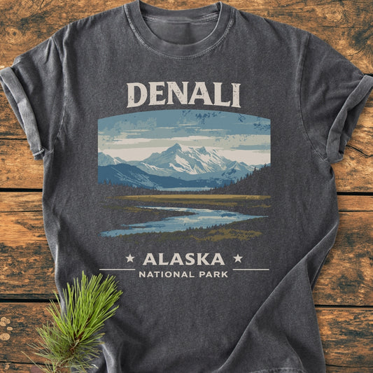 Denali National Park - Vintage Wash Tee featuring snow-capped mountains and forest graphic, laid on rustic wood. Made of soft, garment-dyed 100% cotton, highlighting its relaxed fit and durability.