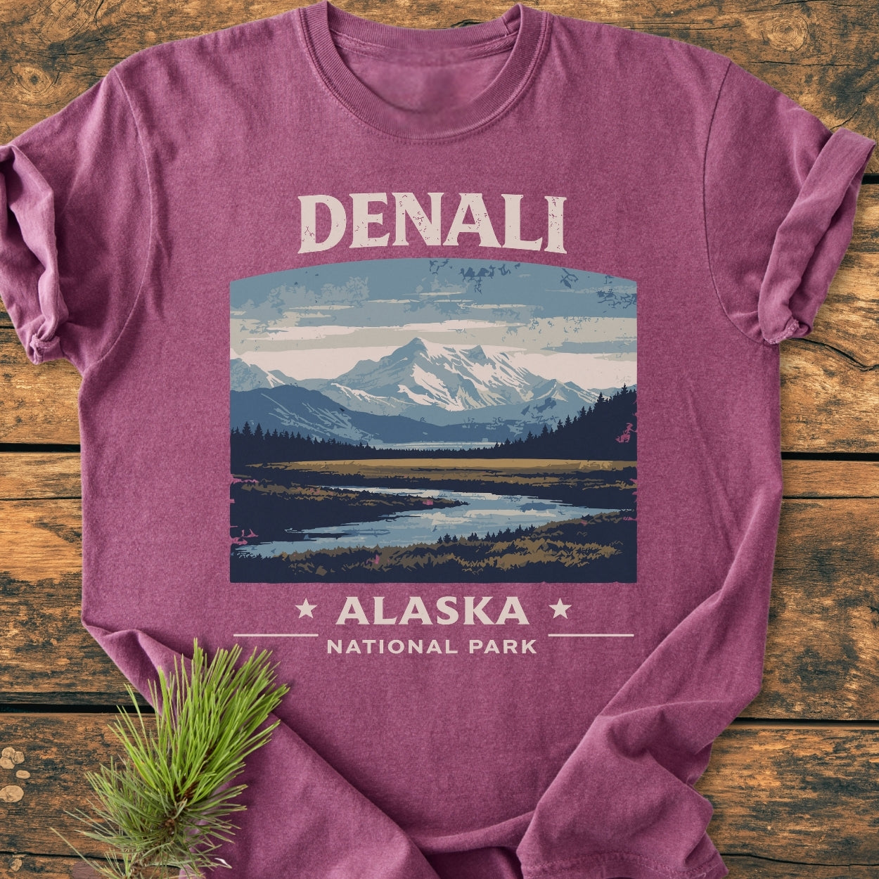 Denali National Park - Vintage Wash Tee: Heather-purple T-shirt featuring a vintage mountain landscape with “DENALI” and “ALASKA NATIONAL PARK” text, laid on a wooden surface with a pine sprig.
