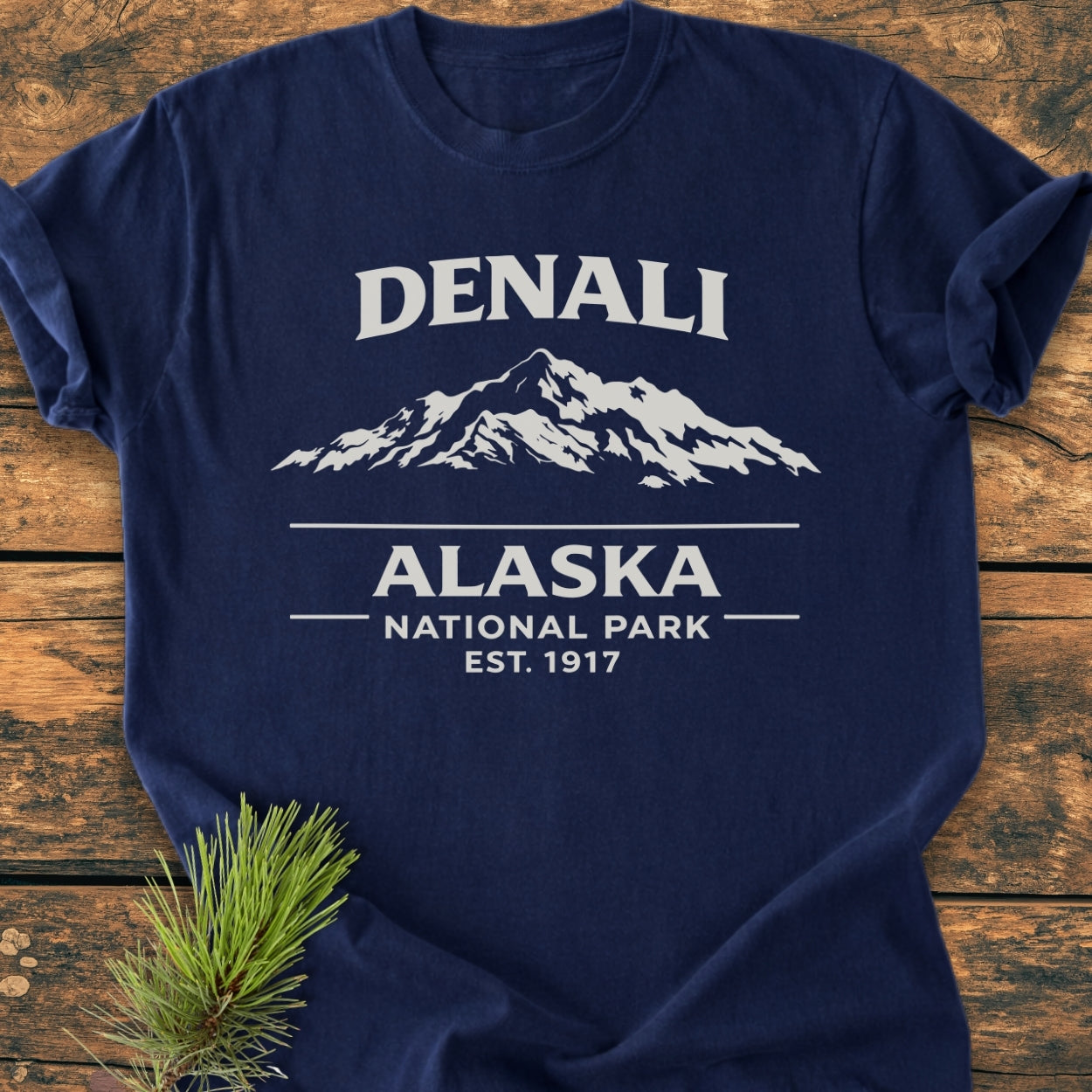 Denali Alaska National Park - Vintage Wash Tee displaying a white mountain graphic and text on a navy base, laid flat on wood with pine needles nearby. Perfectly combines style with comfort and durability.