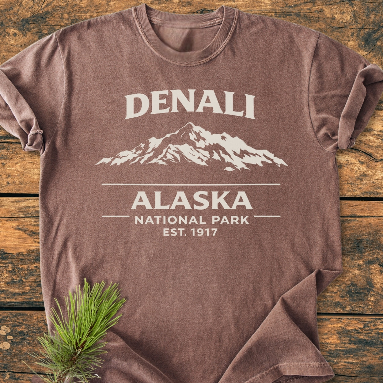 Denali Alaska National Park - Vintage Wash Tee, featuring a mountain silhouette and establishment date, laid on rustic wood with a pine sprig. Made of soft, garment-dyed ring-spun cotton for comfort.