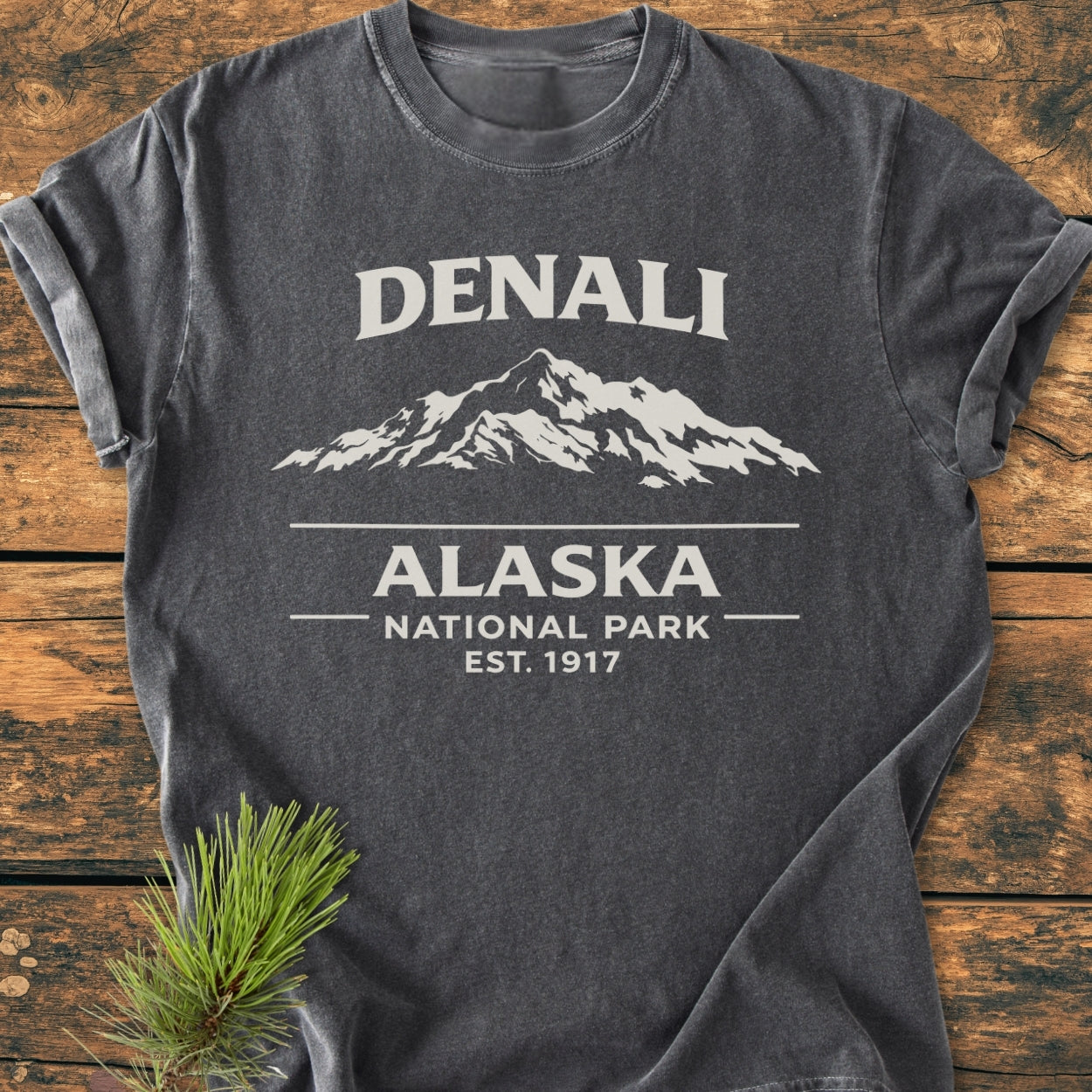 Denali Alaska National Park - Vintage Wash Tee, heather-gray with off-white mountain graphic and text, laid on rustic wood with pine sprig. Made from soft, durable, garment-dyed, 100% ring-spun cotton.