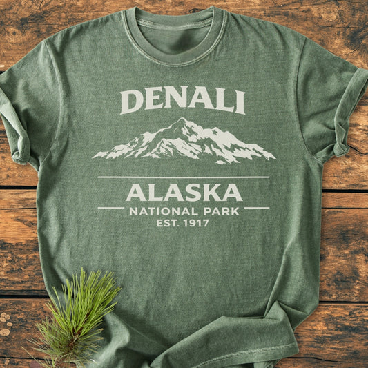 Denali Alaska National Park - Vintage Wash Tee displayed on rustic wood, featuring a stylized mountain graphic and text. A pine branch accents the heather-green, soft-washed, 100% ring-spun cotton fabric.