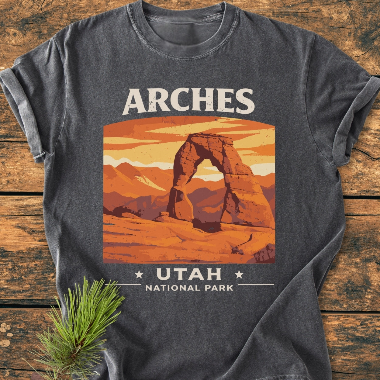 Arches Utah National Park - Vintage Wash Tee: Charcoal-gray T-shirt with Delicate Arch illustration, ARCHES above, UTAH • NATIONAL PARK below; soft-washed, relaxed fit, laid on wooden surface with pine sprig.