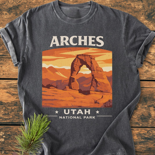 Arches Utah National Park - Vintage Wash Tee: Charcoal-gray T-shirt with Delicate Arch illustration, ARCHES above, UTAH • NATIONAL PARK below; soft-washed, relaxed fit, laid on wooden surface with pine sprig.