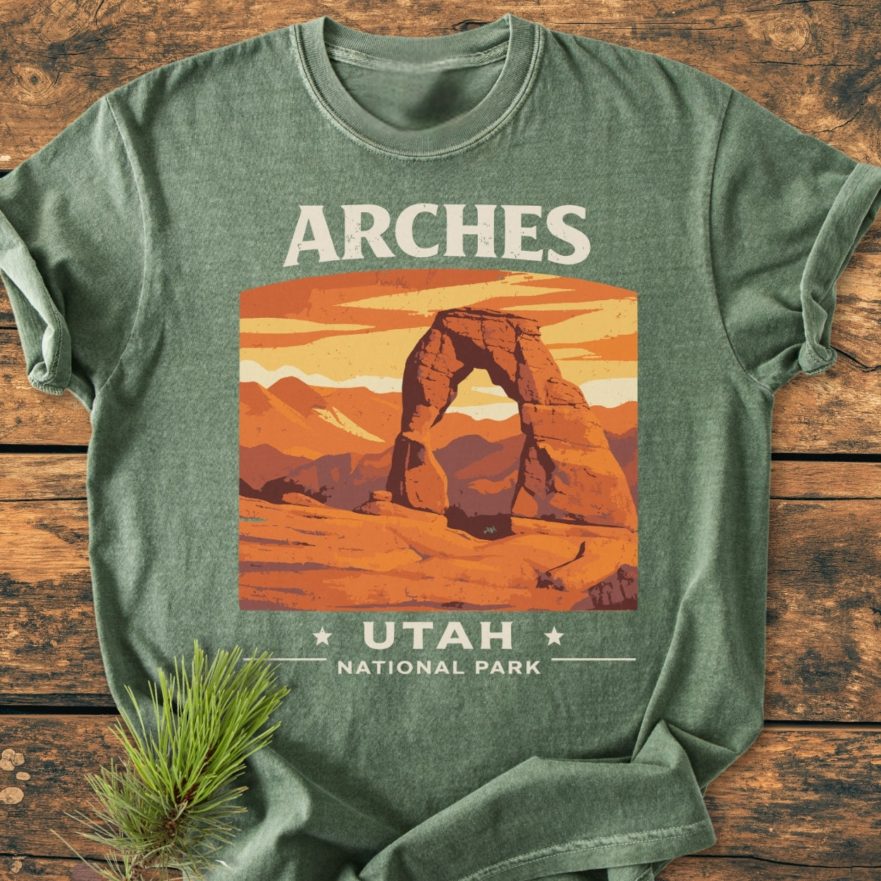 Arches Utah National Park - Vintage Wash Tee in heather-green, featuring a stylized Delicate Arch graphic, displayed flat with a pine bough on a wooden surface, highlighting its relaxed fit and durable design.