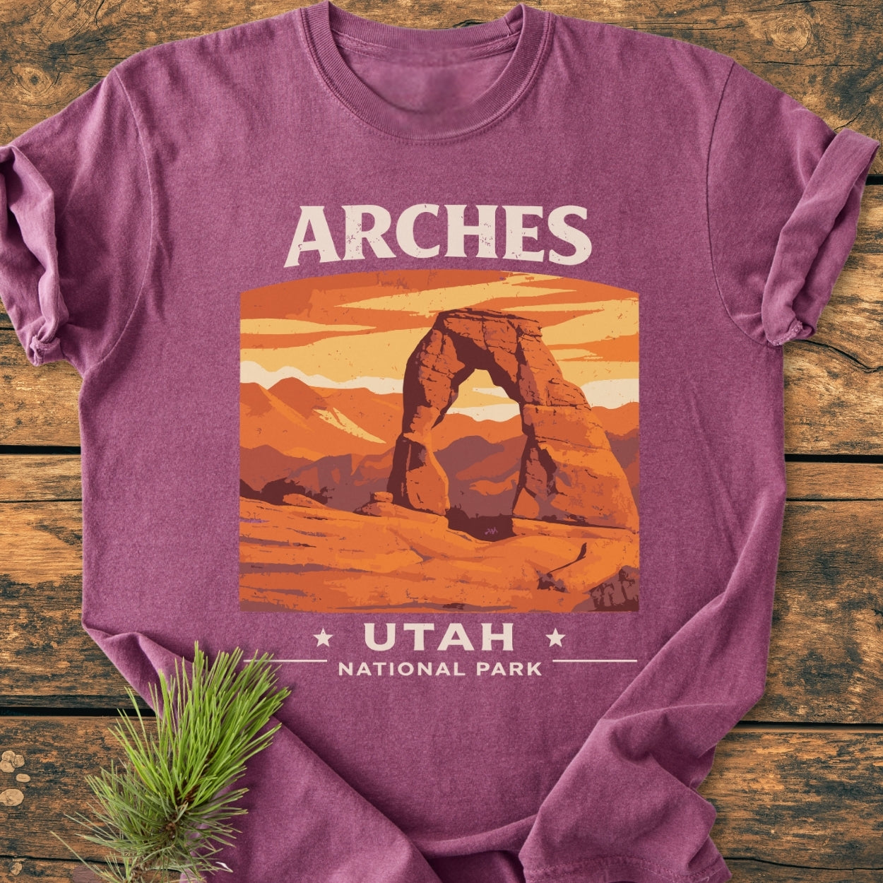 Arches Utah National Park - Vintage Wash Tee folded on wooden surface, featuring a retro Delicate Arch graphic against an orange sunset. Soft-washed, relaxed fit, 100% ring-spun cotton, emphasizing comfort and durability.