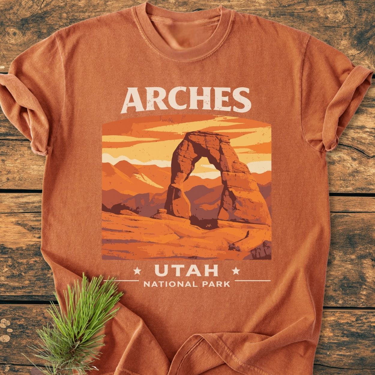 Arches Utah National Park - Vintage Wash Tee, rust-orange with Delicate Arch graphic, laid flat on wood. Features stylized desert scene and ARCHES UTAH NATIONAL PARK text, showcasing soft, garment-dyed cotton fabric.