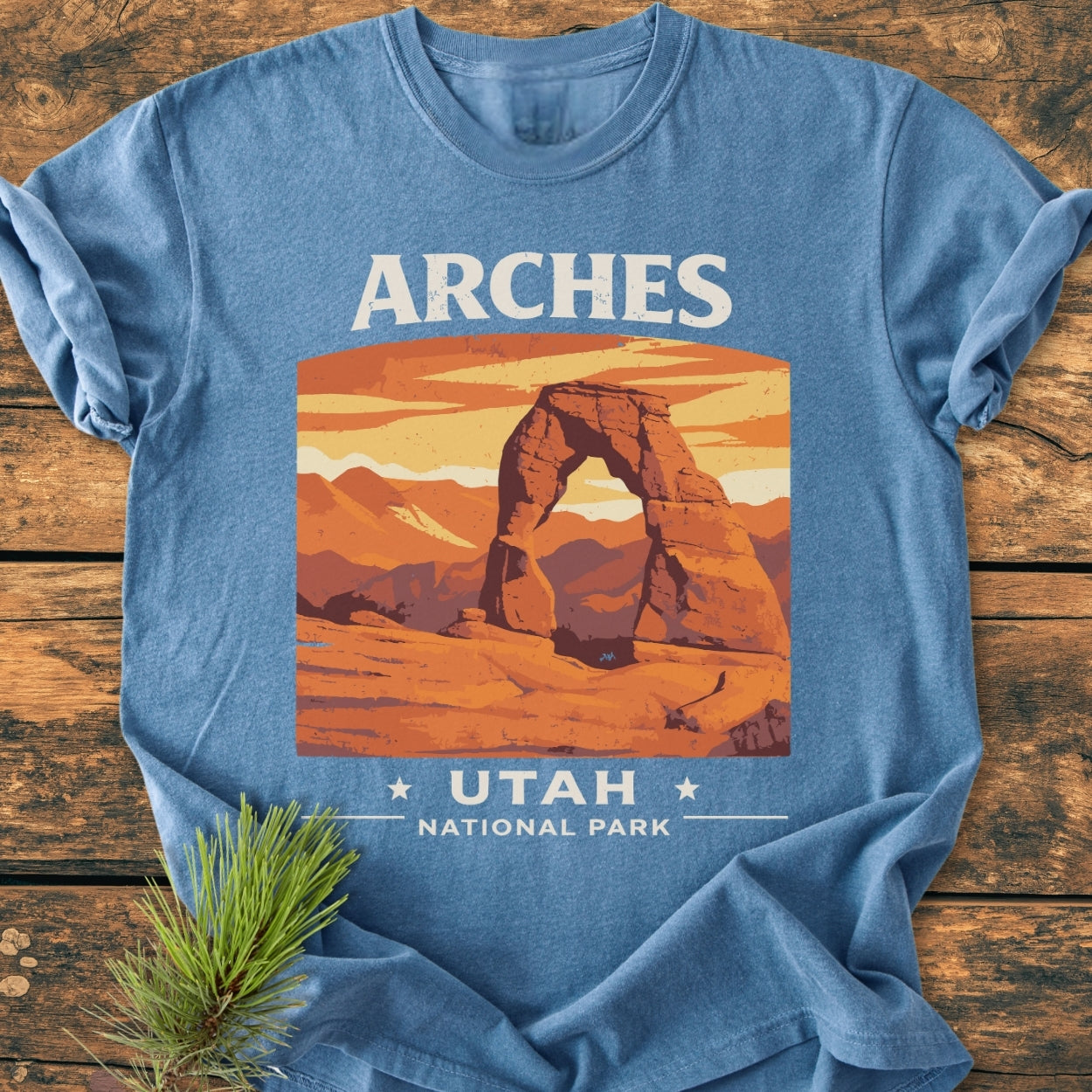 Arches Utah National Park - Vintage Wash Tee featuring retro travel graphic of Delicate Arch against an orange-yellow sky. Displayed on wood with a pine sprig, showcasing its soft, relaxed fit.