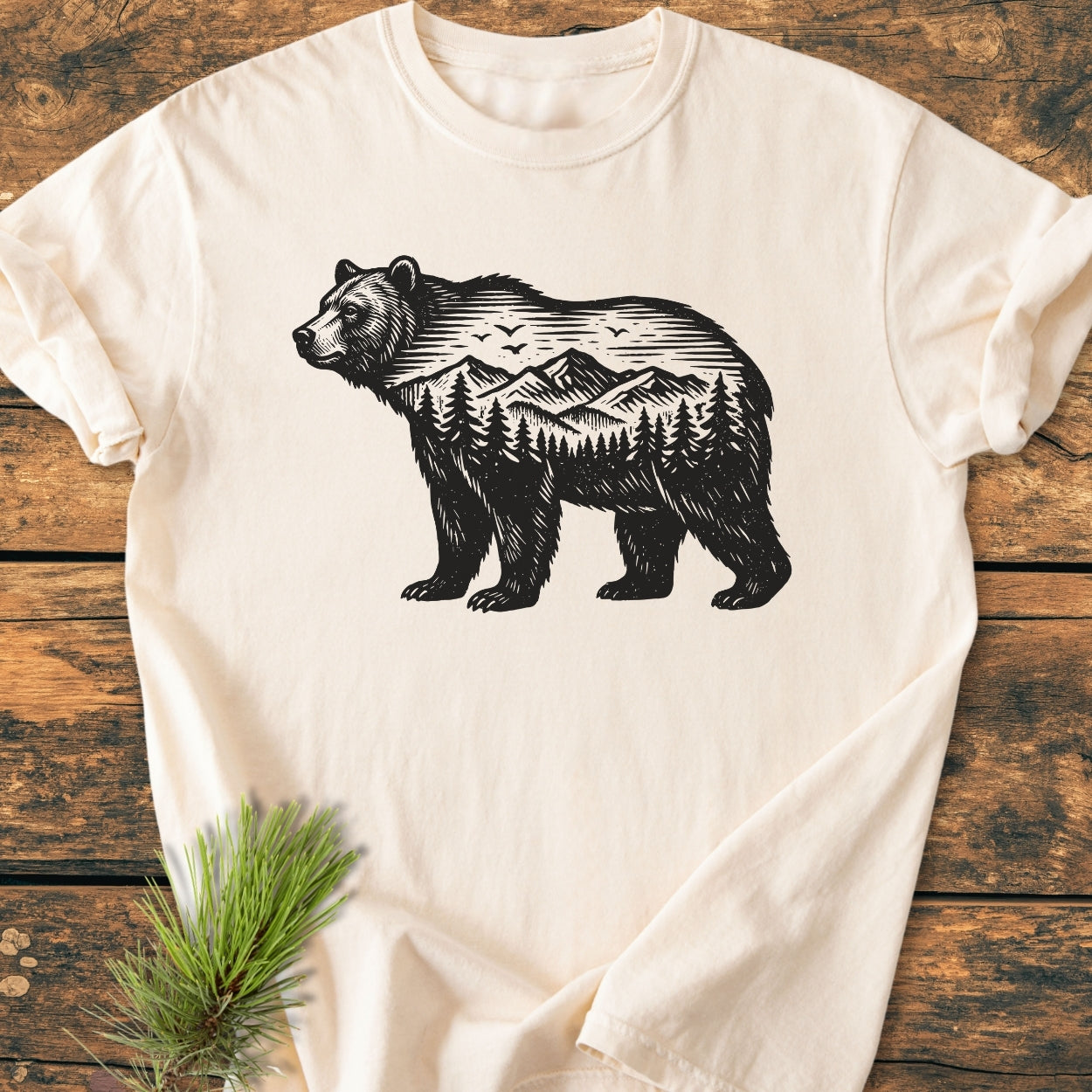The Wilderness Within - Vintage Wash Tee features a black bear silhouette with mountains and trees, displayed on a wooden surface beside a fir sprig, highlighting its unique design and soft texture.