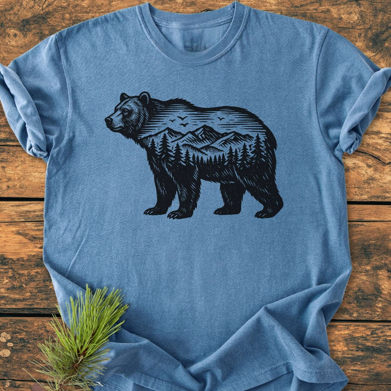The Wilderness Within - Vintage Wash Tee features a bear silhouette with a mountain landscape, crafted from soft-washed, garment-dyed cotton, displayed on a wooden surface with a pine needle sprig.