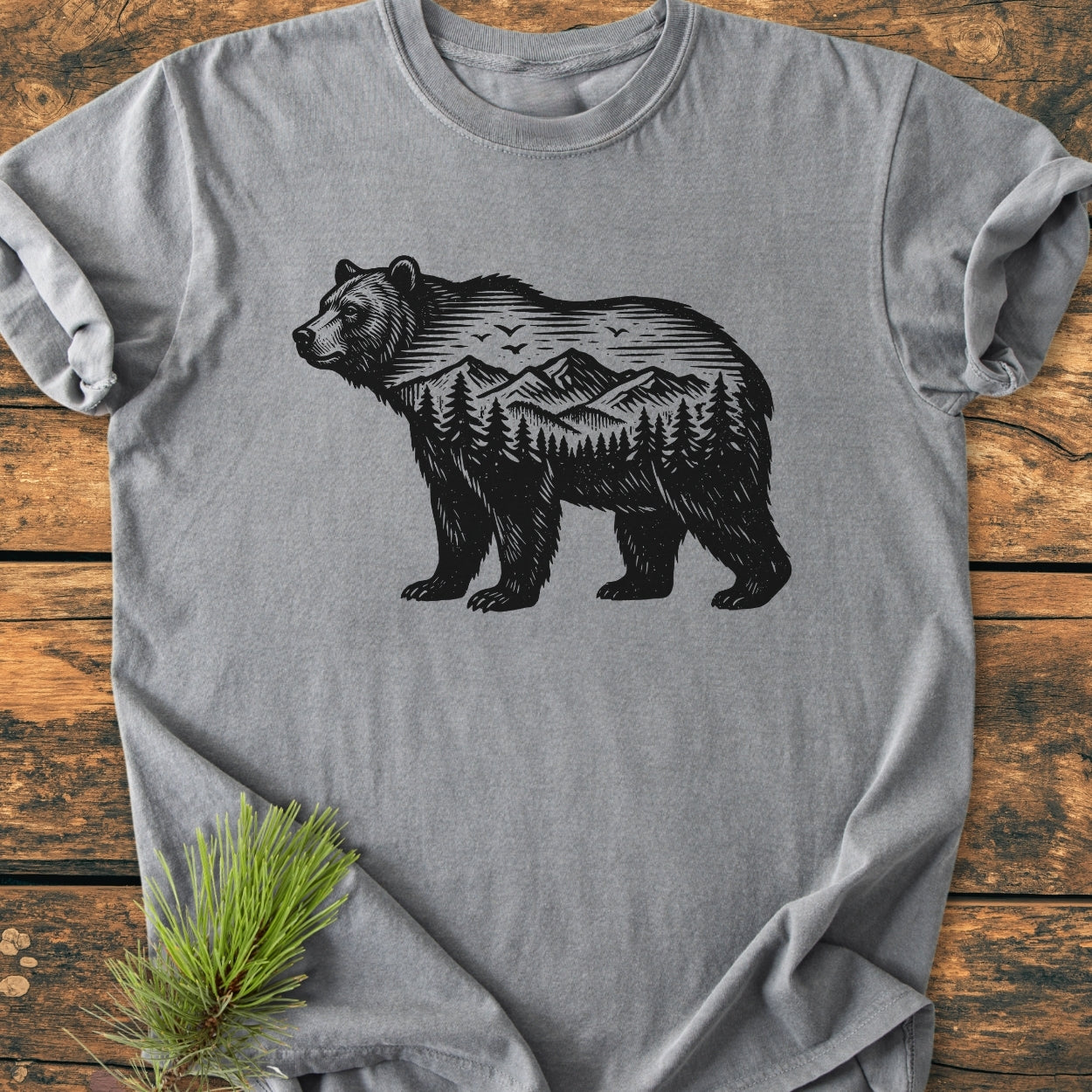 The Wilderness Within - Vintage Wash Tee features a black bear silhouette with mountain and pine designs, lying on rustic wood. A pine sprig adds a natural touch to this heather-gray shirt.