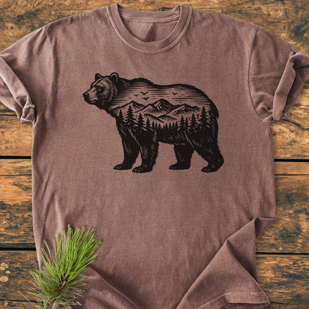 The Wilderness Within - Vintage Wash Tee features a bear silhouette with a mountain scene, laid on a wooden surface with a pine branch accent, showcasing its rustic charm and cozy fit.