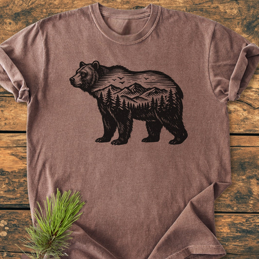 The Wilderness Within - Vintage Wash Tee features a bear silhouette with a mountain scene, laid on a wooden surface with a pine branch accent, showcasing its rustic charm and cozy fit.