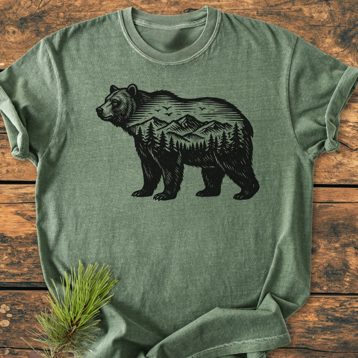 The Wilderness Within - Vintage Wash Tee features a bear silhouette with a mountain landscape, laid on wood, complemented by a pine sprig, emphasizing its outdoorsy style and comfort.