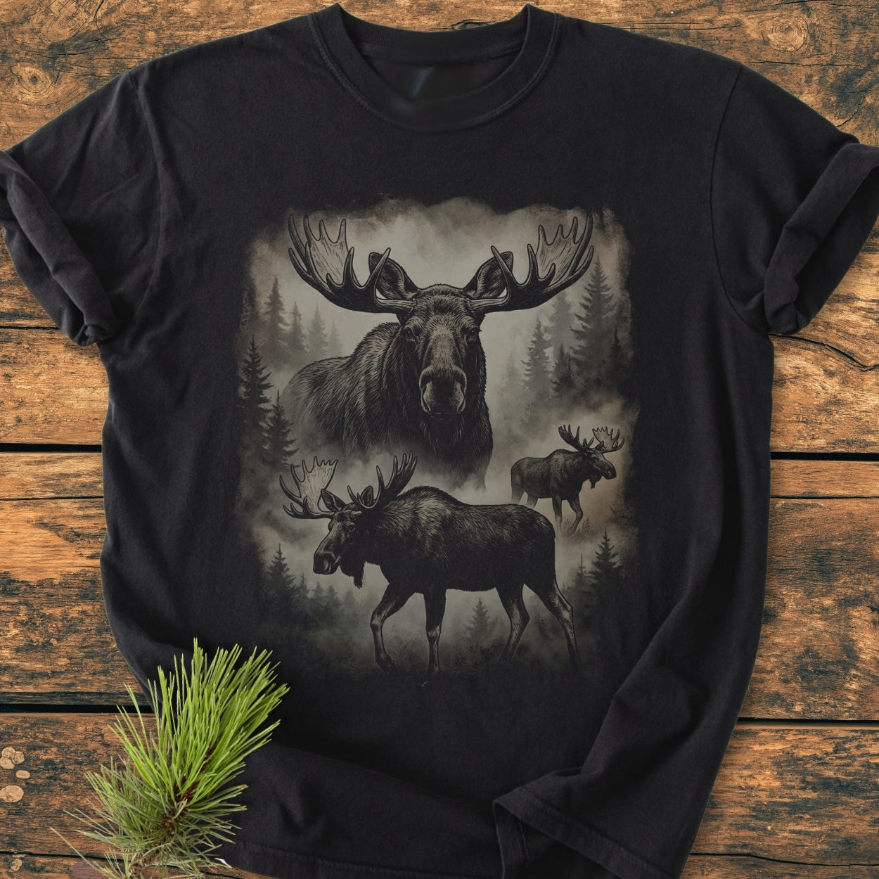 Shadow Moose - Vintage Wash Tee, featuring a forest scene with three moose against misty pine trees on a black T-shirt, laid flat on a wooden surface with a pine sprig nearby.