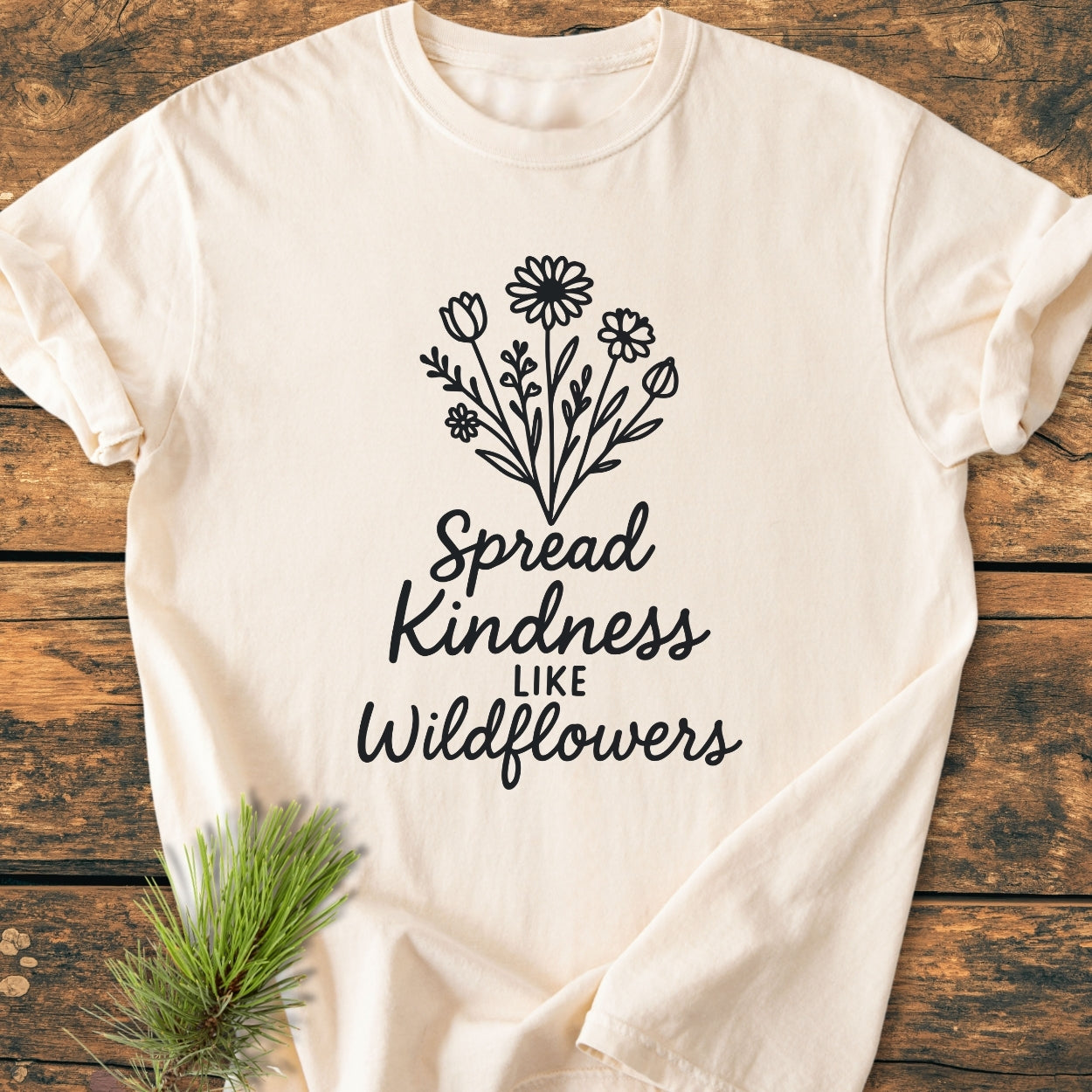 Spread Kindness - Vintage Wash Tee features a central line-drawing of wildflowers with the phrase “Spread Kindness Like Wildflowers” on an off-white tee, laid flat on a wooden surface.