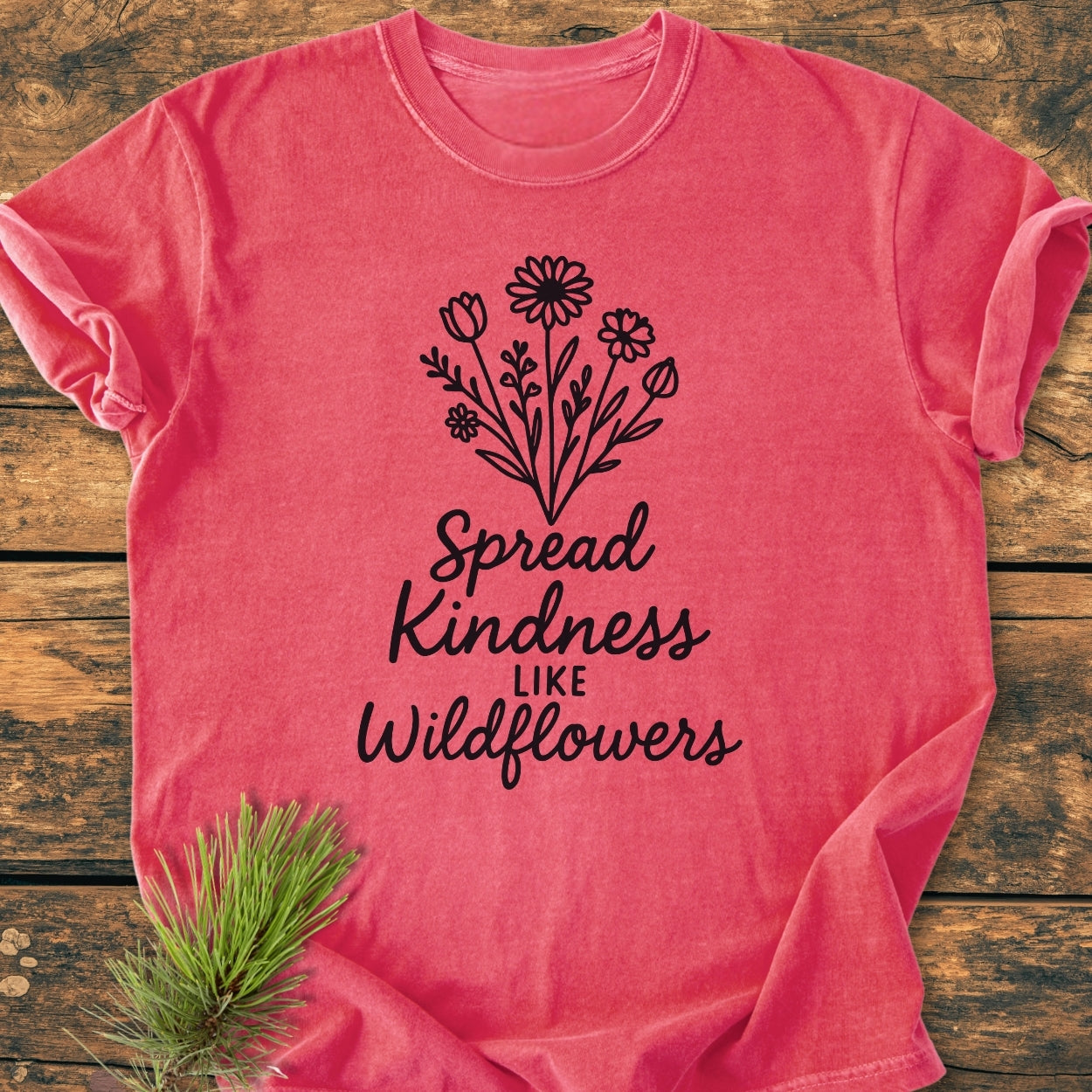 Spread Kindness - Vintage Wash Tee features a black line-drawing of wildflowers with the slogan “Spread Kindness like Wildflowers” on a pinkish-red, garment-dyed T-shirt, laid flat on wood.
