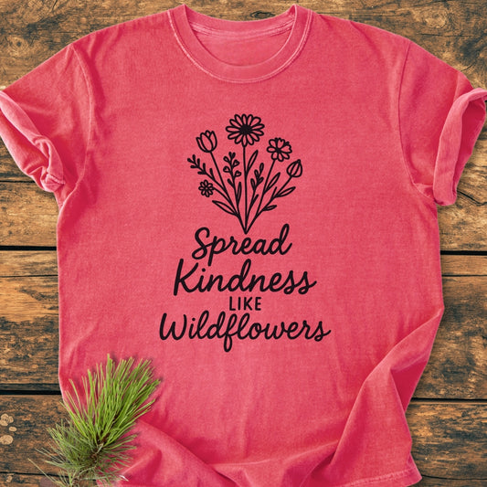 Spread Kindness - Vintage Wash Tee features a black line-drawing of wildflowers with the slogan “Spread Kindness like Wildflowers” on a pinkish-red, garment-dyed T-shirt, laid flat on wood.